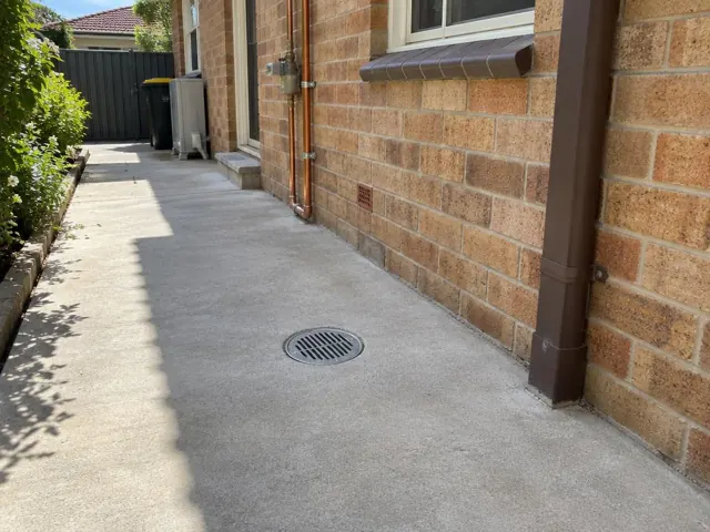 Outdoor grated drain set flush in a dry paved side passage beside a brick house in Rockdale