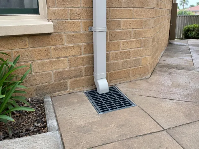 Stormwater grate beside a brick house wall and downpipe on a paved driveway in Brighton-Le-Sands
